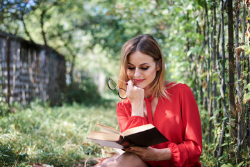 Fototapeta premium Young pretty blond woman, wearing red shirt, sitting by fence in garden holding book and eyeglasses. Close-up picture of student, studying reading book thinking in park in summer. Campus leisure.