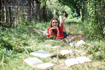 Naklejka na ściany i meble Young blond woman, wearing red shirt, lying on green grass with books in front of her, Student reading book in park forest. Leisure in campus. Reading book addict. Books on the ground,