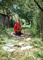Naklejka na ściany i meble Young blond woman, wearing red shirt, sitting on green grass with books in front of her, Student reading book in park forest. Leisure in campus. Reading book addict. Books on the ground,