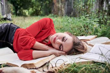 Young blond woman, wearing red shirt, lying sleeping on brown blanket. Creative close-up portrait, with person surrounded with many books. College student, tired on green grass after studying.