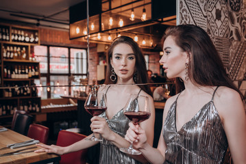 a beautiful young girl in a shiny dress with long hair stands at the mirror with a glass of red wine in her hand