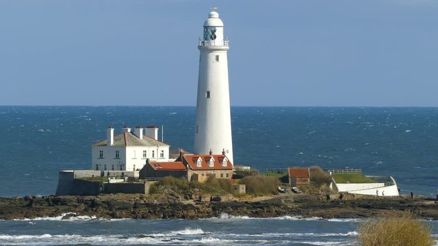 Sunlit lighthouse and properties against a blue sky, with unidentifiable visitors walking around. A tidal causeway links St Mary&rsquo;s Lighthouse back to the nearby shore at Whitley Bay, England, UK.
