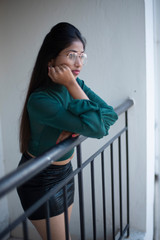 Fototapeta na wymiar Fashion portrait of an young and attractive Indian Bengali brunette girl with green cropped top, skirt and spectacles standing on a balcony in white background. Indian fashion portrait and lifestyle.