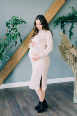 a young beautiful pregnant woman in a beige dress stands in a cozy room and holds her stomach