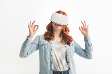 smiling redhead teenager showing ok signs and using vr headset, isolated on white