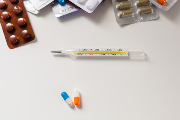 A large number of drugs on a white background next to a thermometer and two capsules of orange and blue
