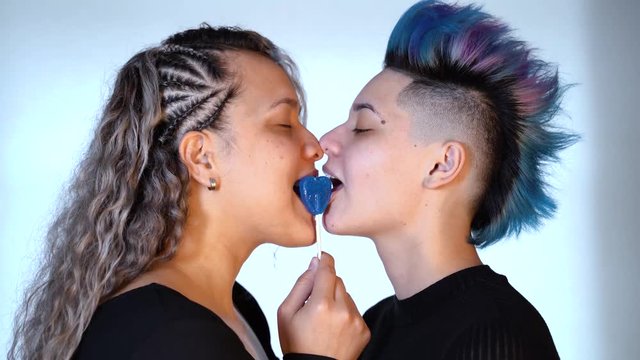 Ethnic homosexual girlfriend playing with heart shaped lollipop to kiss a beautiful punk woman with blue mohawk hairdo. Modern gay love. Lgbt and pride concept. Gay lifestyle. Real lesbian couple.