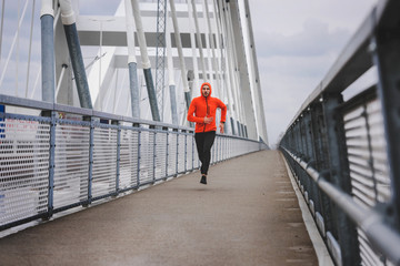 Handsome young athlete running fast along big modern bridge in orange windbreaker jacket. Exercising, Jogging, Sport, Winter. Male athlete running.