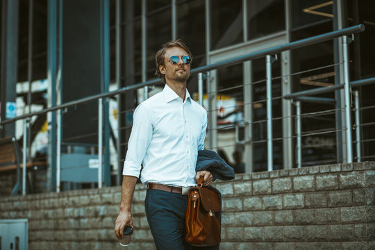 Handsome businessman in white shirt and jacket on his hand walking to work. Young man holds a dispoable cup of coffe in one hand and briefcase in other.