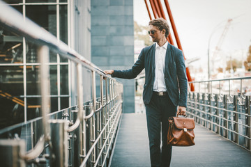 Handsome businessman standing on a bridge of business center. Young trainee in blue sute, sunglasses and briefcase in had on the way to work.