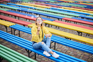 Fototapeta na wymiar Young blond woman, wearing yellow hoody, blue jeans and eyeglasses, sitting on colorful bench in city urban park in summer. Portrait of pretty girl, blowing making soap bubbles, laughing, smiling.