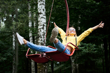 Young blond woman, wearing yellow hoody and blue jeans, riding on carrousel, smiling, laughing in park with green trees in summer. Entertainment in amusement park. Sunday leisure time.