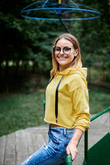 Young blond woman, wearing yellow hoody, blue jeans and eyeglasses, standing on old carrousel in park in summer Three-quarter portrait of girl spending free time in city forest. Summer entertainment.