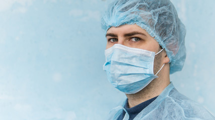 Close up portrait of young european doctor in medical mask. He is looking at camera and isolated on blue background. Coronavirus theme.