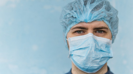 Portrait of young doctor in blue medical mask and cap. Close up. Man is looking at camera. He is isolated on blue background. Coronavirus theme.