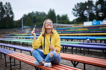 Fototapeta premium Young blond woman, wearing yellow hoody, blue jeans and eyeglasses, sitting on colorful bench in city urban park in summer. Portrait of pretty girl, blowing making soap bubbles, laughing, smiling.