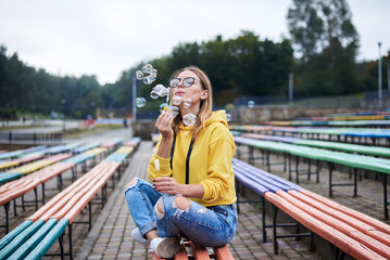 Young blond woman, wearing yellow hoody, blue jeans and eyeglasses, sitting on colorful bench in city urban park in summer. Portrait of pretty girl, blowing making soap bubbles, laughing, smiling.