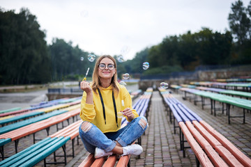 Young blond woman, wearing yellow hoody, blue jeans and eyeglasses, sitting on colorful bench in city urban park in summer. Portrait of pretty girl, blowing making soap bubbles, laughing, smiling.