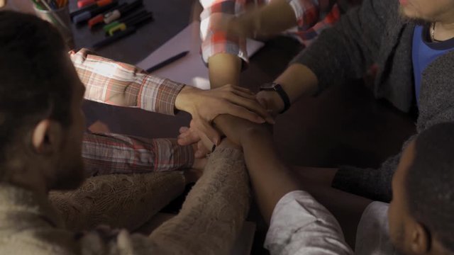 A group of young startupers or freelancers is sitting at a table in a trendy office. Young businessmen put hands of each other's hand and chear up. Unity and teamwork concept.