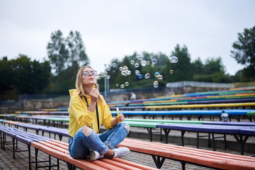 Young blond woman, wearing yellow hoody, blue jeans and eyeglasses, sitting on colorful bench in city urban park in summer. Portrait of pretty girl, blowing making soap bubbles, laughing, smiling.