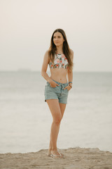 Fototapeta na wymiar An attractive girl with long hair relaxing on the rocky shore of the red sea in a swim top and denim shorts on the sunrise