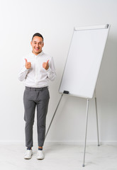 Young latin coaching man with a whiteboard isolated raising both thumbs up, smiling and confident.