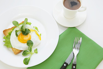 fried egg with a yellow yolk and green salad on a white plate and a cup of black coffee. Table setting with green napkin, fork and knife on a white table. Tasty and healthy breakfast. Top view.