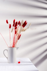 Ceramic mug with dry soft autumn red and white flowers Lagurus Ovatus grass on a table covered textile cloth against wall with shadows from jalousie.