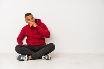 Young latin man sitting on the floor isolated thoughtful looking to a copy space covering mouth with hand.