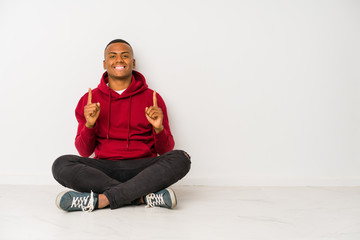 Young latin man sitting on the floor isolated indicates with both fore fingers up showing a blank space.