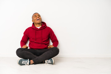 Young latin man sitting on the floor isolated relaxed and happy laughing, neck stretched showing teeth.