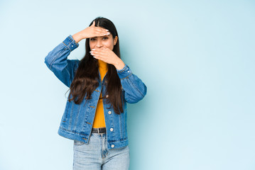 Young indian woman isolated on blue background blink at the camera through fingers, embarrassed covering face.