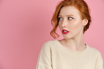 Fototapeta premium A beautiful red-haired girl with red lips in a white sweater stands on a pink background and, turning her head, looks to the side