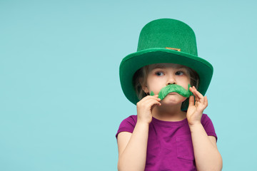 Funny little child girl in green Patricks hat with clover glasses on color background. St. Patricks...