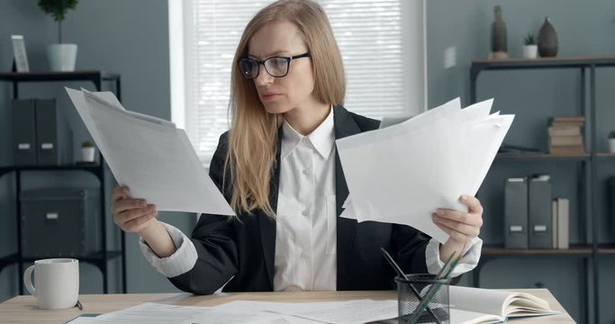Concentrated middle-aged woman in black suit and eyewear sitting at office and searching for important documents among other papers lying on table. Concept of working process and business