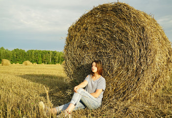 Naklejka na ściany i meble A young girl is sitting near a round stack of straw in a mown field of rye.