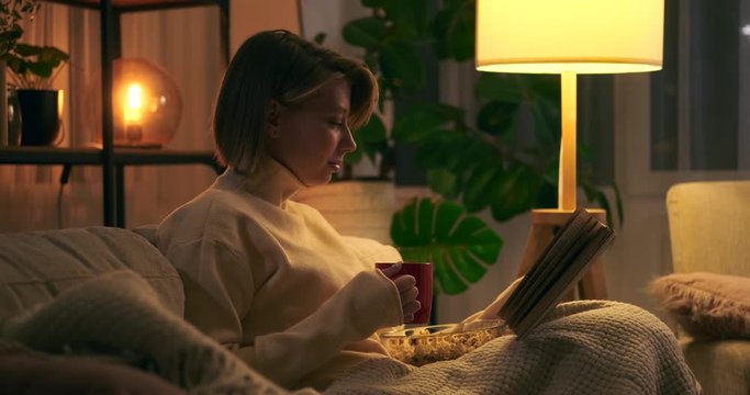 Happy woman drinking coffee and reading book sitting on couch at night