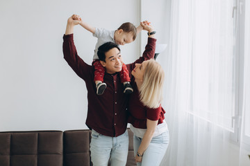 Young asian dad and european blonde mom hold son in her arms in bright room. Multicultural trendy...