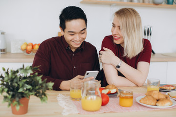 Asian man and his caucasian blonde woman have breakfast. They spend time together in kitchen and look video on the smartphone.