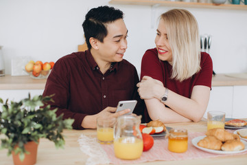 Obraz na płótnie Canvas Asian man and his caucasian blonde woman have breakfast. They spend time together in kitchen and look video on the smartphone.