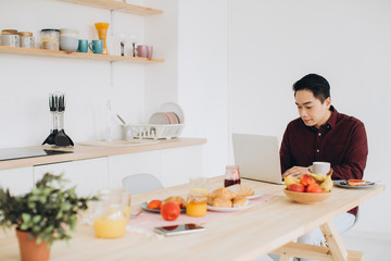 Modern Asian man working on laptop in the kitchen at breakfast.