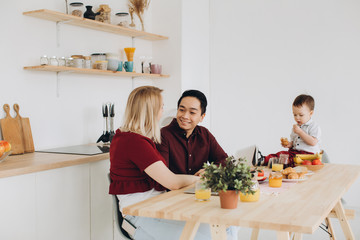 Happy multicultural family. Asian dad and his caucasian blonde wife have breakfast with their beautiful son in the kitchen.