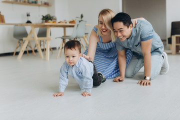 Happy multicultural family having fun together in the kitchen. Asian dad and caucasian mom teach son to crawl