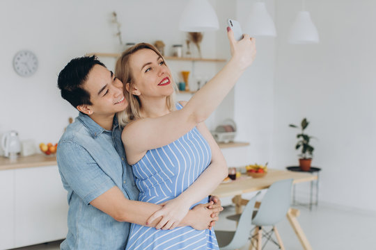 Asian man and his caucasian blonde woman spend time together and making selfie for earch other in the kitchen