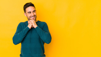 Young caucasian man isolated on yellow bakground keeps hands under chin, is looking happily aside.