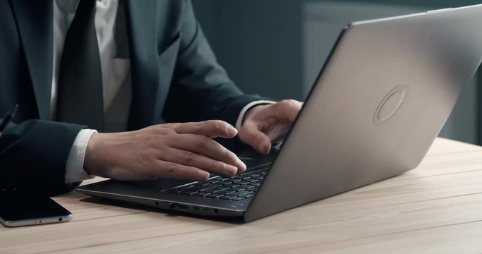 Close up of mature man in black suit sitting at desk and typing on laptop. Successful businessman working on computer at private office.