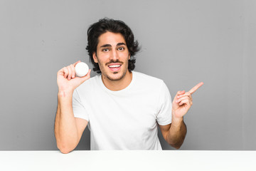 Young man holding a moisturizer smiling cheerfully pointing with forefinger away.