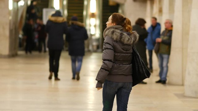 Girl in winter jacket gestures to follow her in the lobby of metro station in Moscow, Russia. Young beautiful woman walks around the station hall  in subway and waves her hand, urging  to come closer.