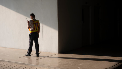 Asian security guard in safety vest walking on sidewalk of parking garage, he using walkie-talkie...