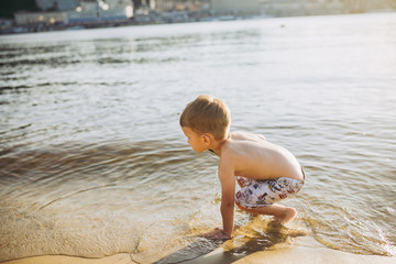 Little Caucasian boy child sits near a river on a sandy beach and touches the surface of the water with his hands. The theme is the flow of time, a short life, the meaning and purpose of existence